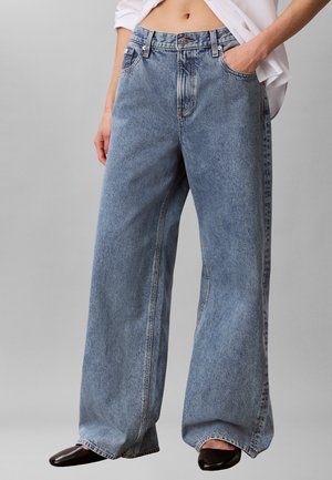 Persona que lleva vaqueros azules de cintura alta y pierna ancha con bolsillos delanteros, combinados con zapatos negros y una camisa blanca parcialmente visible.