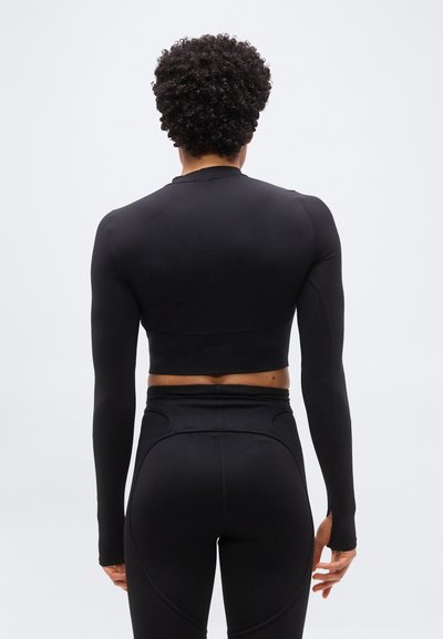 Άτομο με σγουρά μαλλιά που φοράει στενό κοντό μαύρο μπλουζάκι με μακριά μανίκια και μαύρα κολάν ψηλής μέσης, όρθιο με την πλάτη προς την κάμερα.