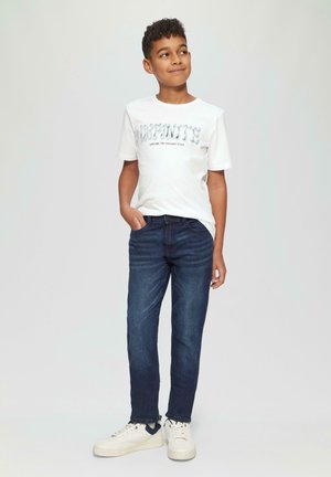 Witte t-shirt met de tekst "INFINITE" in het blauw, gecombineerd met donkerblauwe jeans en witte sneakers met blauwe accenten. Model staat met de handen op de heupen.