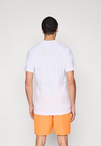 Camiseta deportiva blanca de manga corta con textura sutil, combinada con pantalones cortos de color naranja brillante. Enfoque en material transpirable y ajuste moderno.