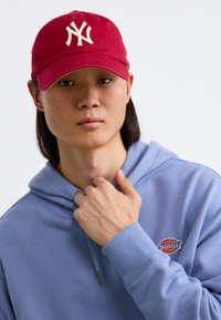 Red cap with white logo, blue hoodie with ribbed cuffs, and small logo patch. Model has long hair, hands near neck. Neutral background.