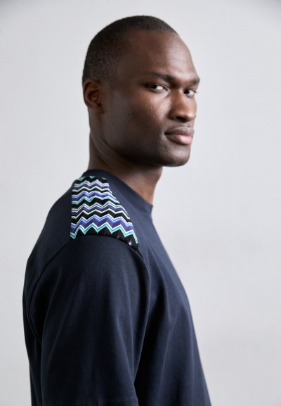 T-shirt bleu marine avec un motif en zigzag texturé et coloré en vert et bleu sur l'épaule. Le tissu semble doux et léger.