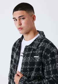 Jonge man met kort haar draagt een diamanten oorbel, zilveren ketting, wit T-shirt met opdruk en zwarte geruite jas, kijkt iets naar links.