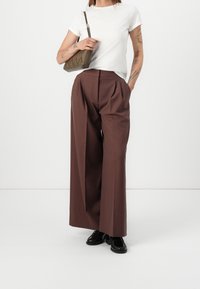 Braune weite Hosen mit vorderen Falten, kombiniert mit einem weißen T-Shirt mit kurzen Ärmeln. Accessoires sind eine strukturierte Handtasche in olivgrün.