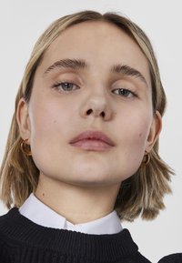 Giovane donna con capelli biondi corti, che indossa orecchini a cerchio dorati, un maglione nero e una camicia bianca con colletto, che guarda con fiducia in avanti.