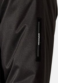 Pantaloni sportivi neri morbidi con una texture liscia e una vestibilità aderente, caratterizzati da una tasca verticale con zip con il logo "EA7" sul lato.