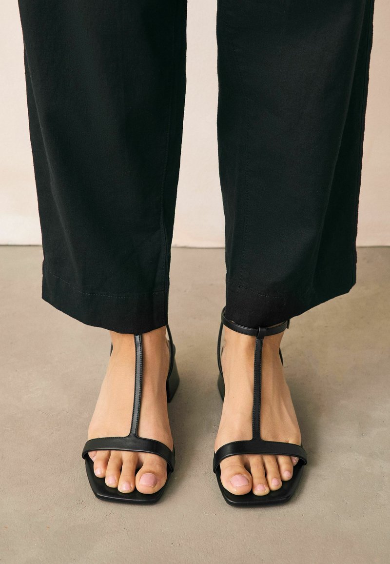 Zwarte leren sandalen met een teenslipperontwerp en vierkante neus, met minimalistische banden en een gladde textuur. Gecombineerd met zwarte wijde broek.