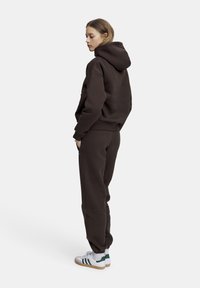 Brun sweatshirt och matchande joggerbyxor, tillverkade av mjukt tyg, med huva, ribbade muddar och elastisk midja. Vita sneakers med gröna detaljer.