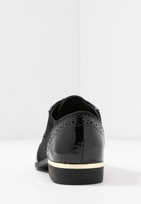 Chaussure noire avec un talon en cuir verni brillant, dessus en daim, détails brogue décoratifs et un talon bas accentué d'or.