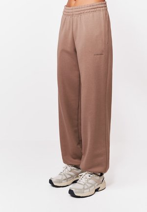 Persoană purtând pantaloni sport maro largi și adidași albi, stând pe un fundal simplu.