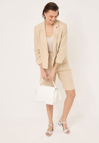 Beige linneblazer och shorts-set, ihopparat med en åtsittande ljus tanktop, vit handväska och grå klacksandaler med blockklack. Släta ytor och enkel design.