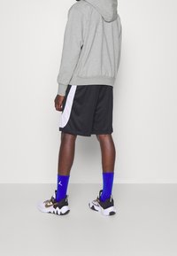 Personne portant un sweat à capuche gris, un short noir et blanc, des chaussettes bleu vif et des chaussures de sport blanches, vue de dos sur un fond uni.