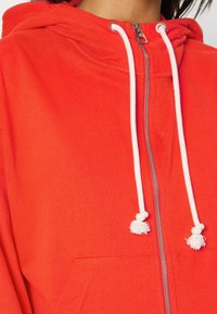 Sudadera con cremallera roja, hecha de un tejido suave, que cuenta con cordones blancos, un bolsillo frontal y un cierre de cremallera suave.