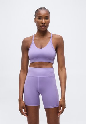 Kvinde iført matchende lavendel sports-bh og høj taljet biker-shorts, stående mod en ensfarvet lys baggrund, med front mod kameraet.