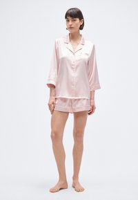 Σετ πυτζάμας από ροζ σατέν που περιλαμβάνει τοπ με κουμπιά και μανίκια τριών τετάρτων και αντίστοιχα σορτς, με λεία υφή και διακριτική λάμψη.