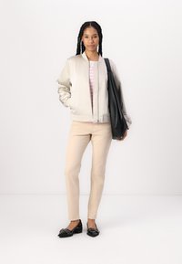 Bomber jacket beige chiaro con una texture liscia, indossato sopra una camicia a righe rosa e bianca. Pantaloni beige a costine e scarpe piatte nere. Borsa nera.
