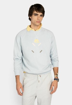 Joven con sudadera azul claro con gráfico de bates de cricket cruzados y gorra amarilla, camisa amarilla con cuello, pantalones a rayas y reloj de pulsera.