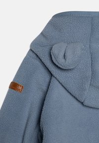 Blå fleecejakke med hette og avrundede øredetaljer samt et brunt skinnmerke nærermed. Myk tekstur og koselig design.