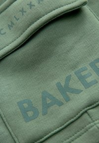 Zelená bavlnená látka s hladkým povrchom, na ktorej je výrazne a trochu vystúpené šedé písmeno "BAKER". Viditeľný detaily šitia.