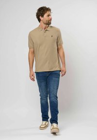 Hombre caminando, viste una camisa polo beige, vaqueros azules y zapatillas beige y blancas, mirando hacia su izquierda contra un fondo blanco.