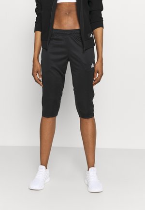 Person iført sorte Adidas cropped træningsbukser, sort lynlåstrøje, hvid sports-bh og hvide sneakers mod en ensfarvet baggrund.