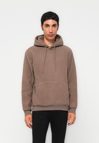 Young man wearing a brown fleece hoodie with front pocket and drawstrings, paired with black pants, standing against a plain light background.