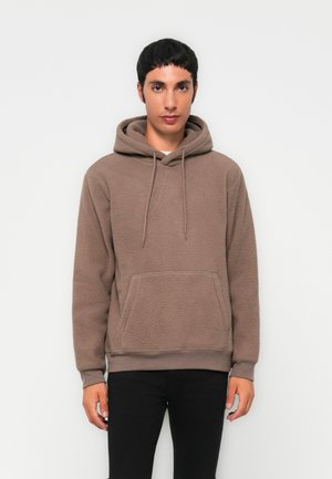 Young man wearing a brown fleece hoodie with front pocket and drawstrings, paired with black pants, standing against a plain light background.