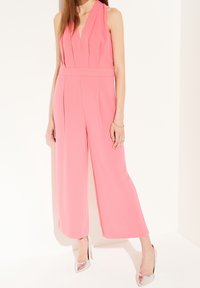 Růžový overal bez rukávů s výstřihem do V, plisovaným designem, širokými nohavicemi a zdůrazněným pasem. Kombinováno se stříbrnými lodičkami na vysokém podpatku.