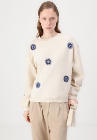 Ljusbeige sweatshirt med marinblå blomsterbroderi. Har rund halsringning, lös passform och ribbade ärmslut. Sammanfogas med ljusbeiga byxor.