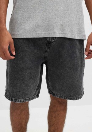 Hombre con pantalones cortos de mezclilla negra desgastada y camiseta gris, de pie con las manos relajadas a los lados.