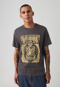 Tricou din bumbac gri, cu un design grafic aurit ce include o vultur, stele și text. Mâneci scurte și guler clasic rotund.
