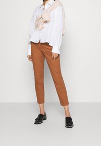 Brązowe spodnie o dopasowanym kroju w połączeniu z białą koszulą zapinaną na guziki. Na ramionach zarzucony jasnoróżowy sweter. Czarne loafersy z srebrnymi akcentami.