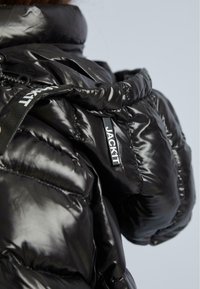 Glänzende schwarze Daunenjacke mit Kapuze, die genähte Details und einen schwarz-weißen Reißverschluss mit "JACKIT"-Markierung zeigt.