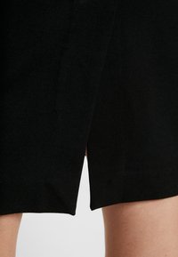 Närbild av en svart kjol med en framslits, som visar en del av bärarens lår mot en enkel bakgrund.