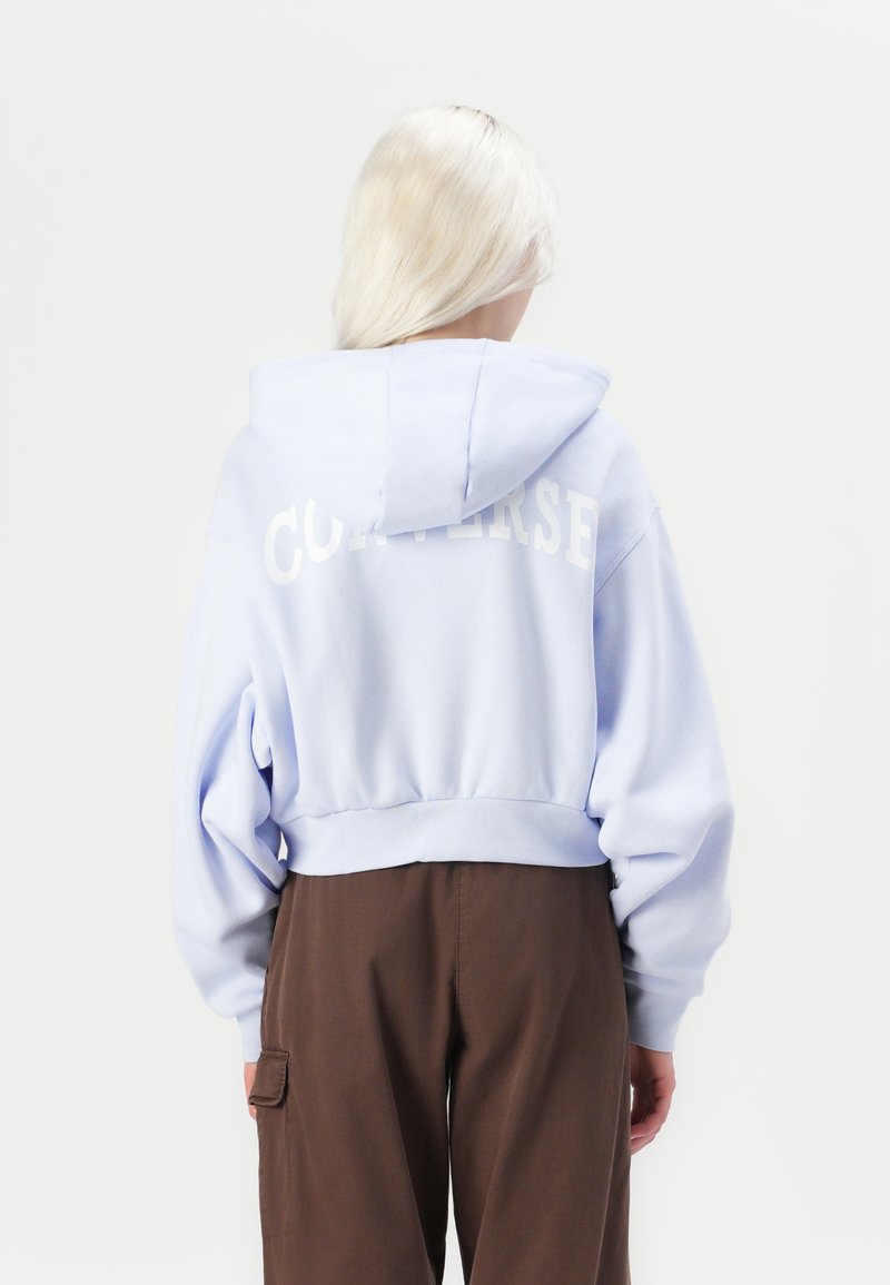 Sudadera de corte corto en un suave color azul claro, con un gran logotipo blanco de "CONVERSE" en la parte posterior. Mangas largas y capucha.