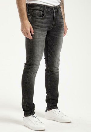 Czarne jeansy slim fit z wyblakłą teksturą, pięć kieszeni i szczegóły przeszyć, w połączeniu z białymi sneakersami w neutralnym otoczeniu.