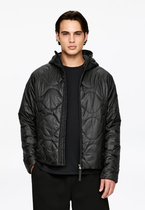 Jeune homme aux cheveux foncés portant une veste matelassée noire à capuche sur une chemise noire, debout devant un fond blanc uni.