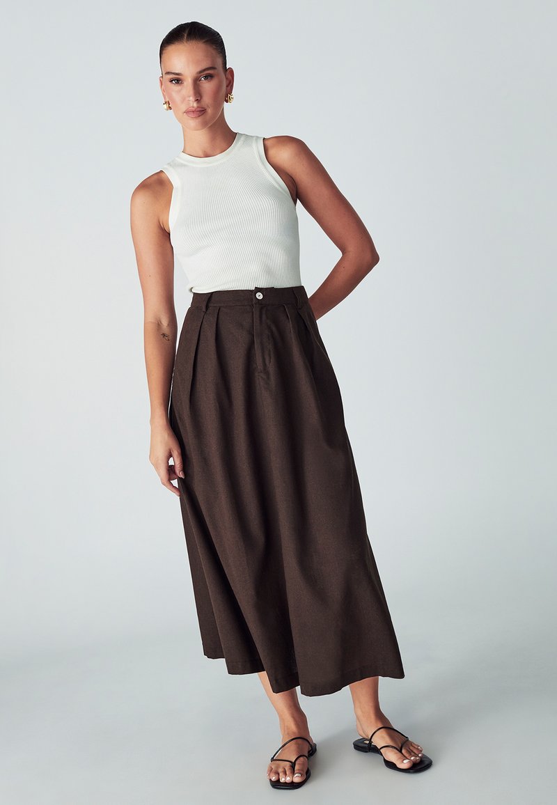Weißes, geripptes Tanktop kombiniert mit einem langen, dunkelbraunen, plissierten Rock mit einer Knopfleiste vorne. Schwarze flache Sandalen runden das Outfit ab.