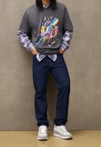 Graues Grafik-T-Shirt, über einem karierten Langarmshirt getragen, kombiniert mit dunkelblauen Jeans und weißen Sneakers mit grauen Akzenten.
