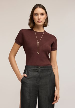 Kobieta w teksturowanym brązowym krótkim rękawie, ciemnych spodniach z wysokim stanem i długim złotym naszyjniku, z jedną ręką w kieszeni.