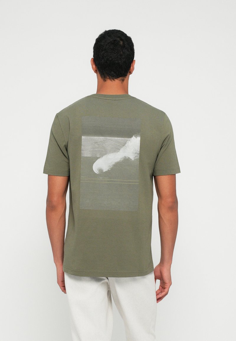 Oliwkowa koszulka z krótkim rękawem wykonana z bawełny, z prostokątnym nadrukiem fali na plecach w odcieniach szarości i bieli.