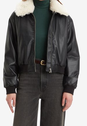 Vrouw draagt een zwarte leren bomberjack met een witte shearling kraag, groene coltrui, zwarte riem en donkergrijze spijkerbroek.