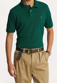 Grön pikétröja med korta ärmar, med en lila logotyp. Matchad med khakifärgade byxor och ett flerfärgat randigt bälte med bruna detaljer.