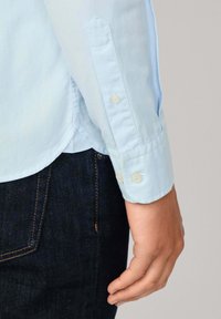 Camicia a maniche lunghe di un blu chiaro, in tessuto di cotone, con polsini abbottonati e orlo curvo. Indossata sopra pantaloni in denim scuro.
