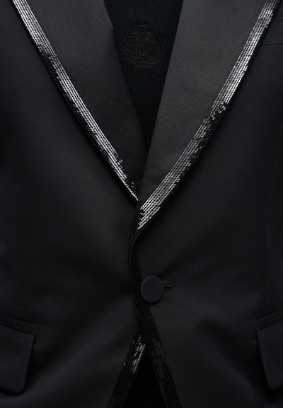 Μαύρο σακάκι σμόκιν με γυαλιστερή διακόσμηση από παγιέτες στο πέτο και ένα μαύρο κουμπί που κλείνει πάνω από σκούρο πουκάμισο με κεντημένο έμβλημα.