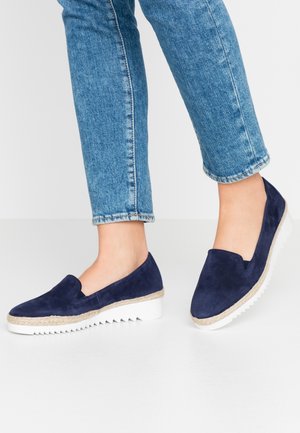 Person iført blå ruskind slip-on-sko med hvide rillede såler og lyseblå denimjeans mod en ensfarvet hvid baggrund.