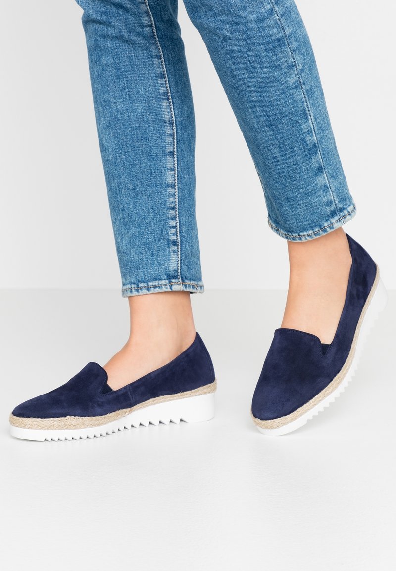 Personne portant des chaussures en daim bleu à enfiler avec des semelles blanches rainurées et un jean en denim bleu clair sur un fond blanc uni.