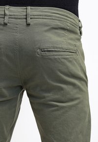 Olivově zelené bavlněné kalhoty s boční kapsou a poutky na opasek, s hladkou texturou a přiléhavým střihem z pohledu zezadu.