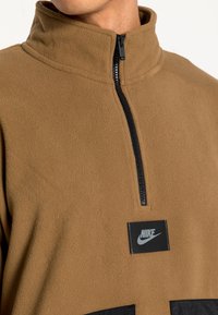 Brun fleece-pulli med zip-kant, med en svart Nike-logotyp och en svart dragkedja. Texturerat tyg och sydda detaljer.