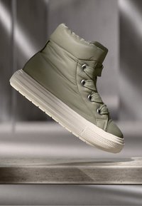 Padded olive green high-top sneaker with a textured upper, round toe, lace-up design, and contrasting cream-colored rubber sole.